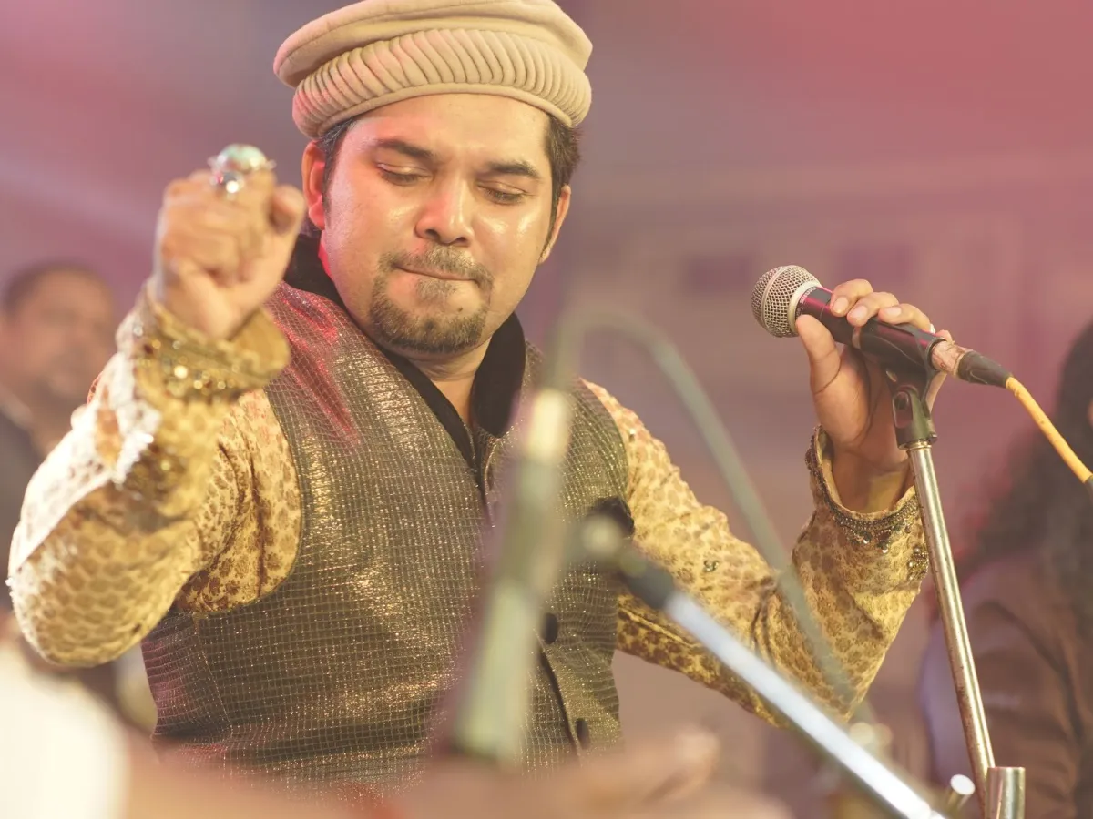 A qawwali singer mid-phrase at an intimate gathering, one hand raised in an expressive gesture, close to the microphone