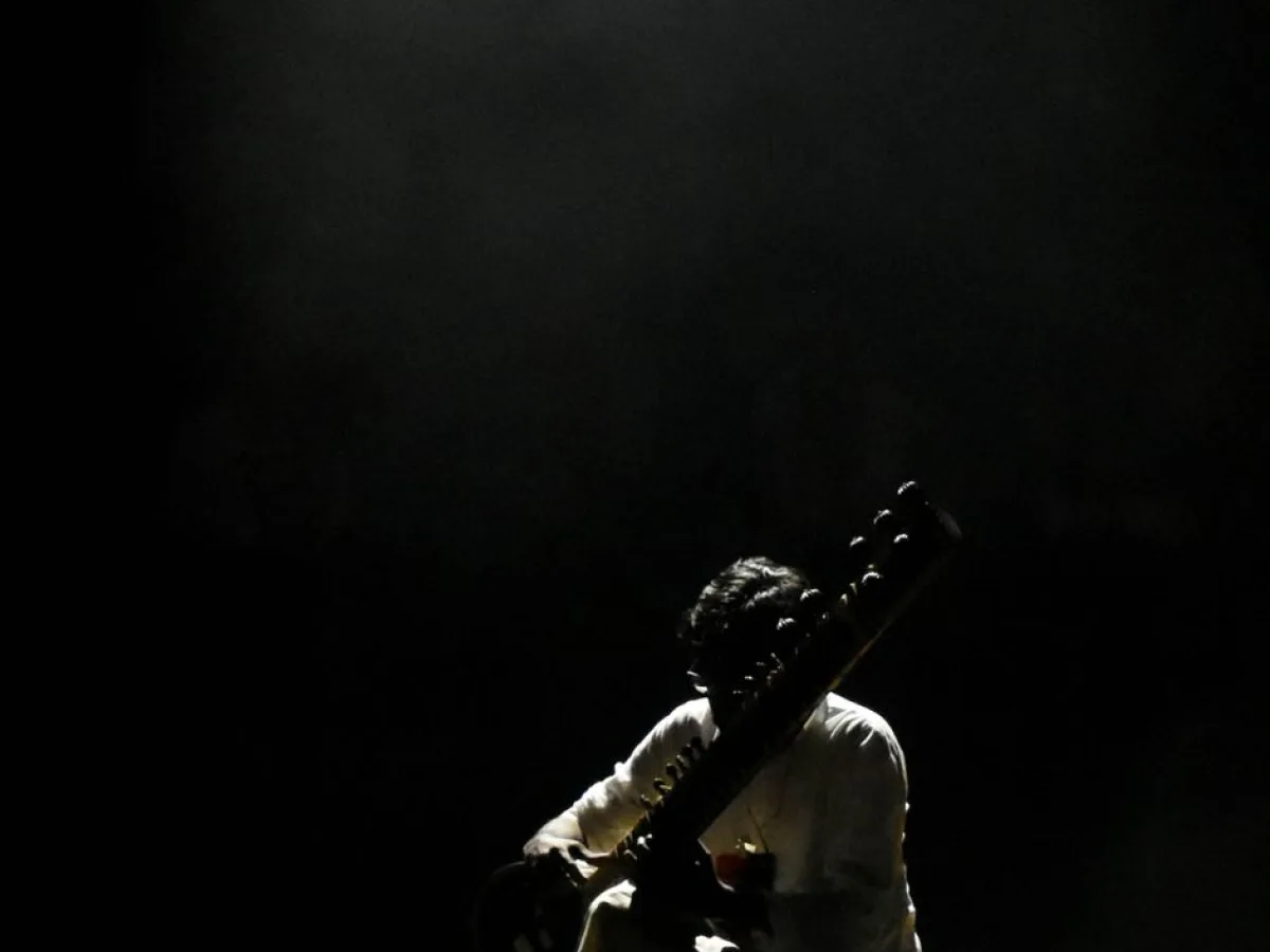 A sitar player silhouetted under a single shaft of light on a darkened stage