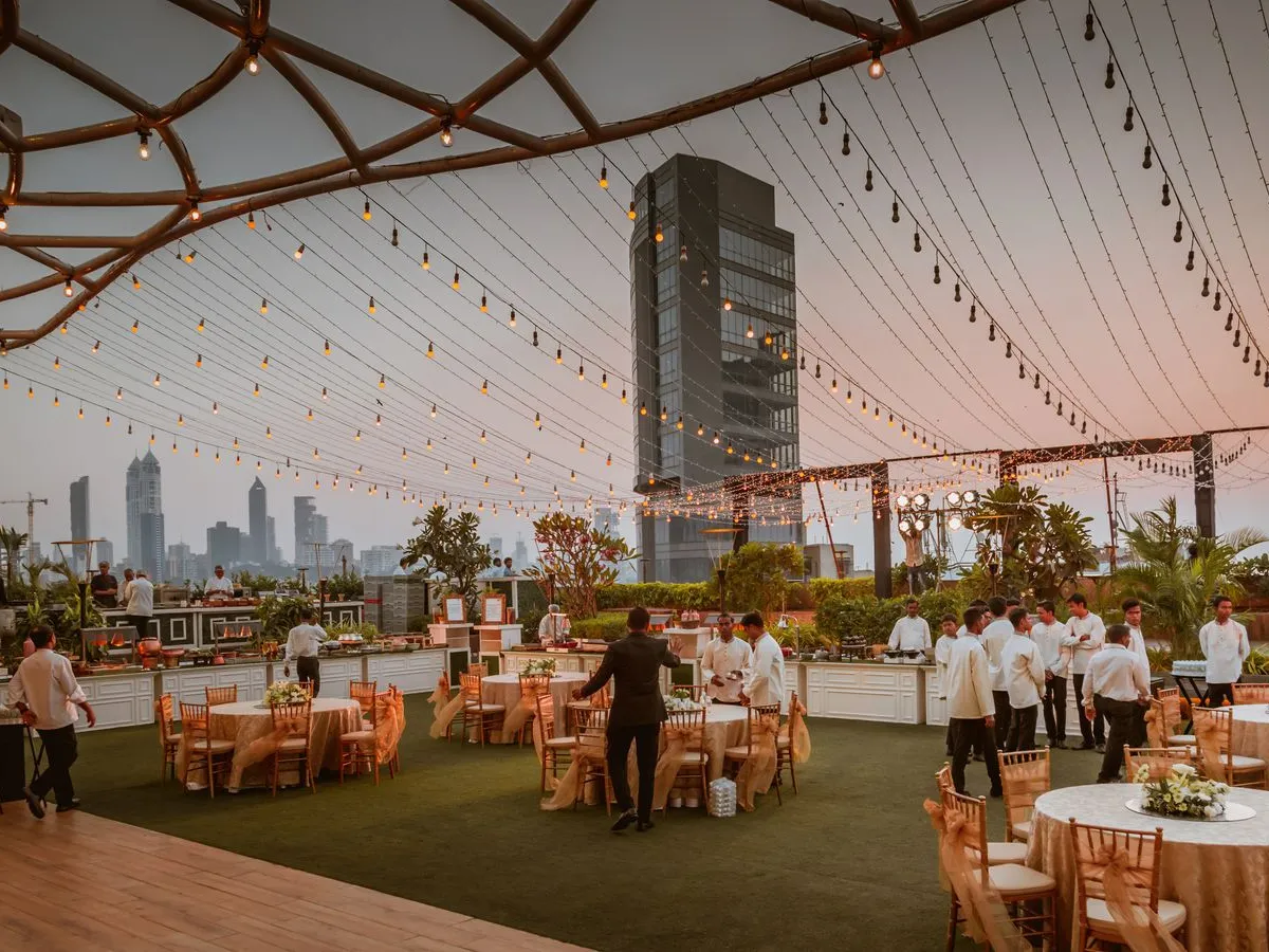 An open-air rooftop venue strung with a canopy of warm lights against the city skyline at dusk